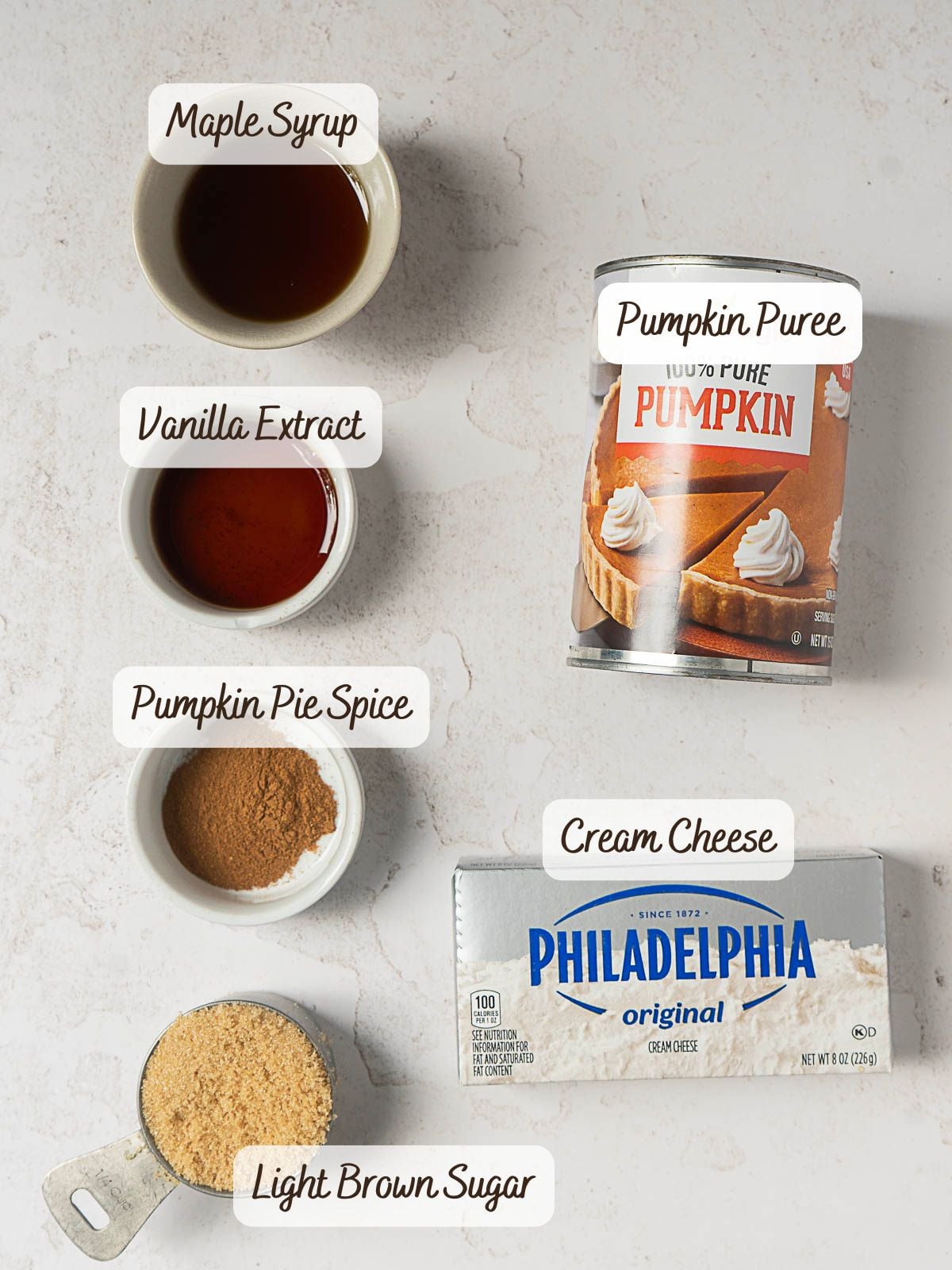 Ingredients on a white surface: a bowl each of maple syrup, vanilla extract, pumpkin pie spice, and light brown sugar, plus a can of pumpkin puree and a block of cream cheese—perfect essentials for whipping up a creamy Pumpkin Pie Dip or any delicious pumpkin dessert.