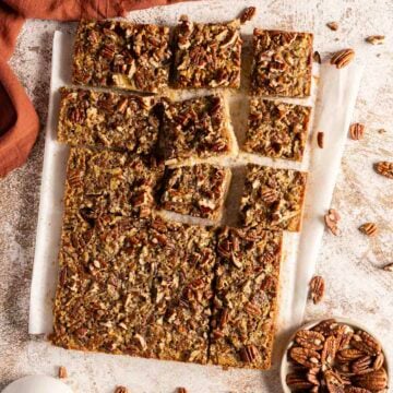 Pecan pie bars cut into squares on parchment paper, with a bowl of whole pecans nearby.