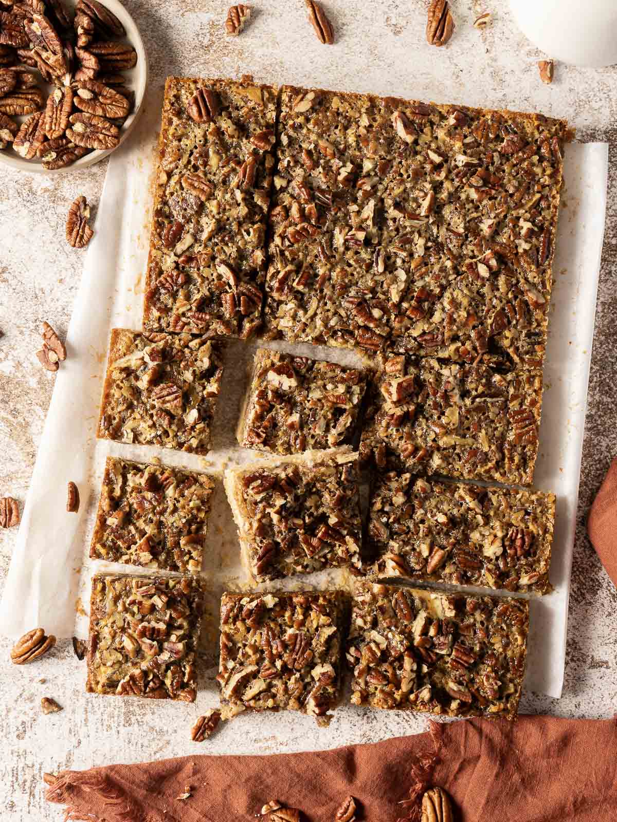 Pecan pie bars cut into squares on parchment paper, with a small bowl of pecans nearby.